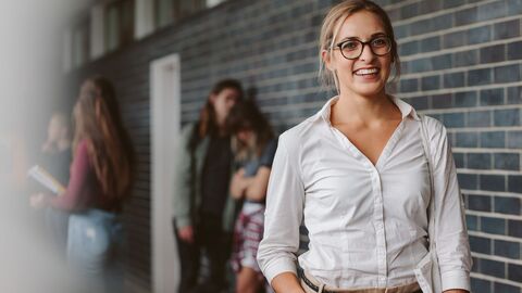 Bild zum Thema Eine Lehrerin steht in einem Flur einer Schule; im Hintergrund stehen Schüler