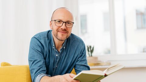 Ein Mann sitzt mit einem Buch auf einem gelben Sofa und schaut den Betrachter an 