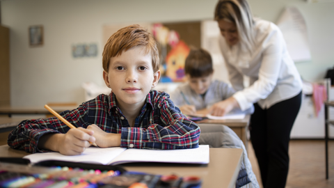 Ein junger Schüler, der im Unterricht sitzt und von seinem Heft hochschaut; im Hintergrund geht die Lehrerin durch die Klasse