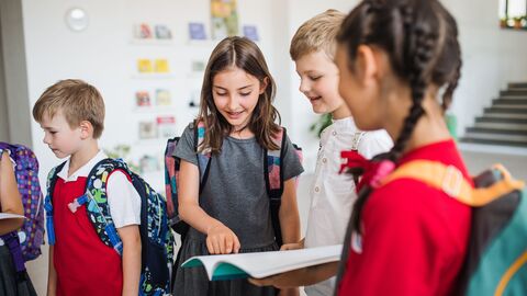 Ein Gruppe Kinder im Gespräch miteinander; sie stehen im Flur einer Schule mit ihren Ranzen auf dem Rücken