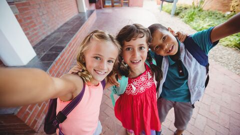 Drei Kinder mit Ranzen, die gerade ein Selfi von sich vor ihrer Schule machen