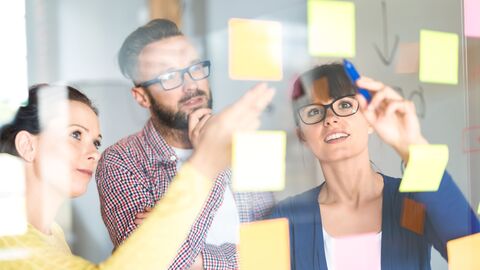 Landesweite Aufgaben Eine Gruppe Mitarbeiter klebt bunte Post-Its an eine Glasscheibe